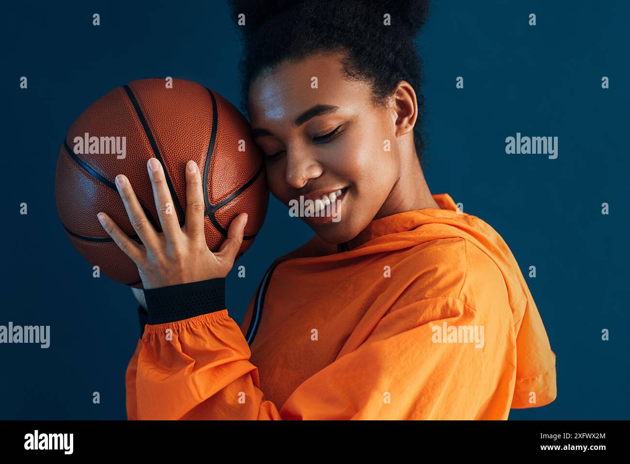 Gros plan d'une jeune femme souriante avec un ballon de basket. Femme avec les yeux fermés portant des vêtements de sport orange debout sur un fond bleu. Banque D'Images