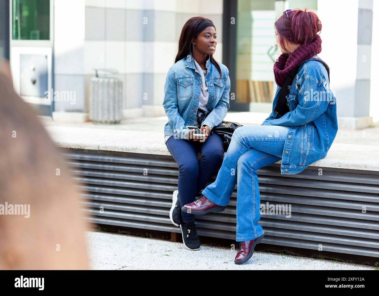 Deux amis, de race blanche et de l'Afrique de l'appartenance ethnique, de parler assis sur un banc à l'extérieur. Banque D'Images
