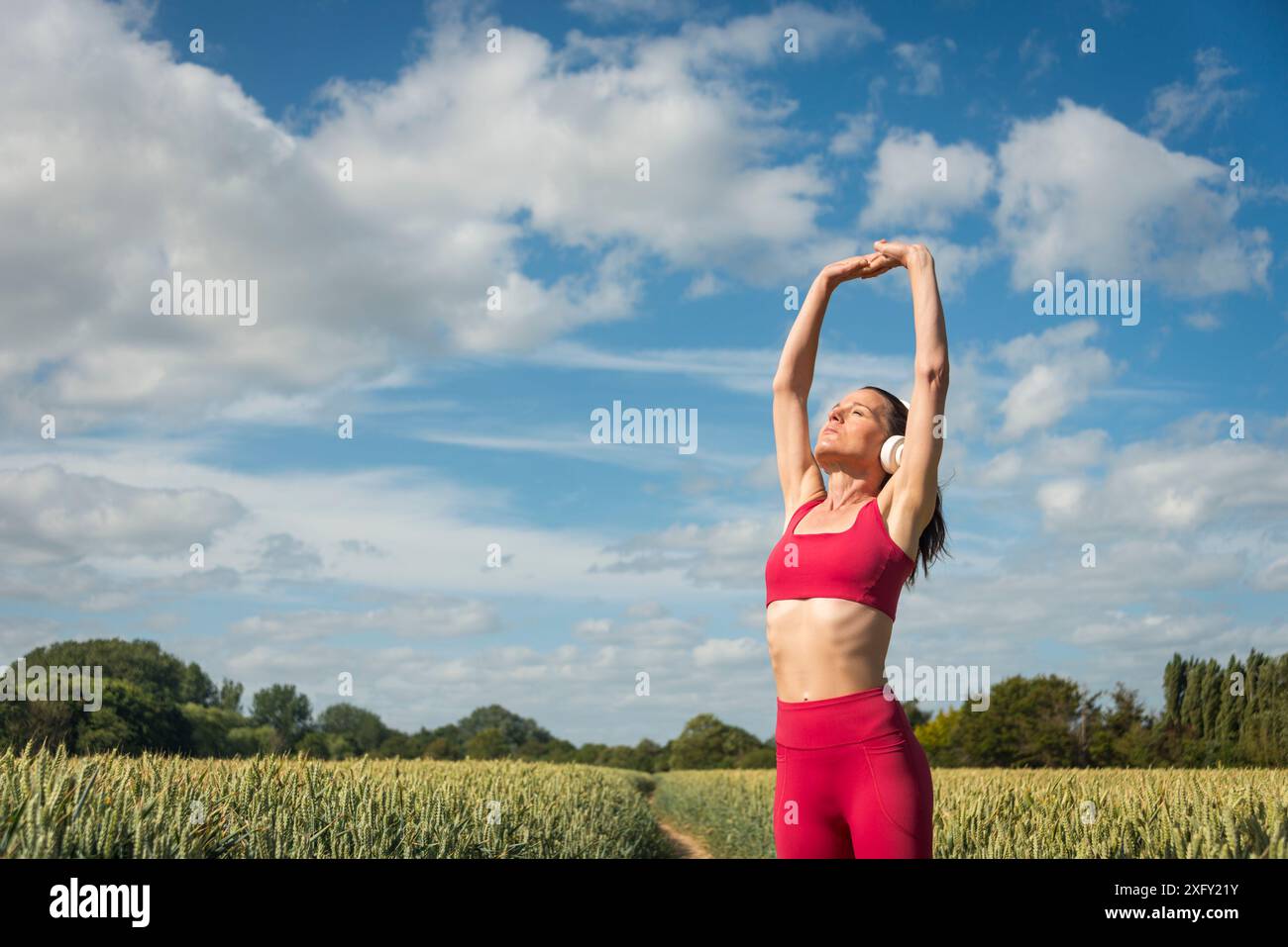 Femme sportive avec les bras levés faisant des exercices d'étirement dans le champ de blé, portant des vêtements de sport rouges avec un ciel bleu. Banque D'Images