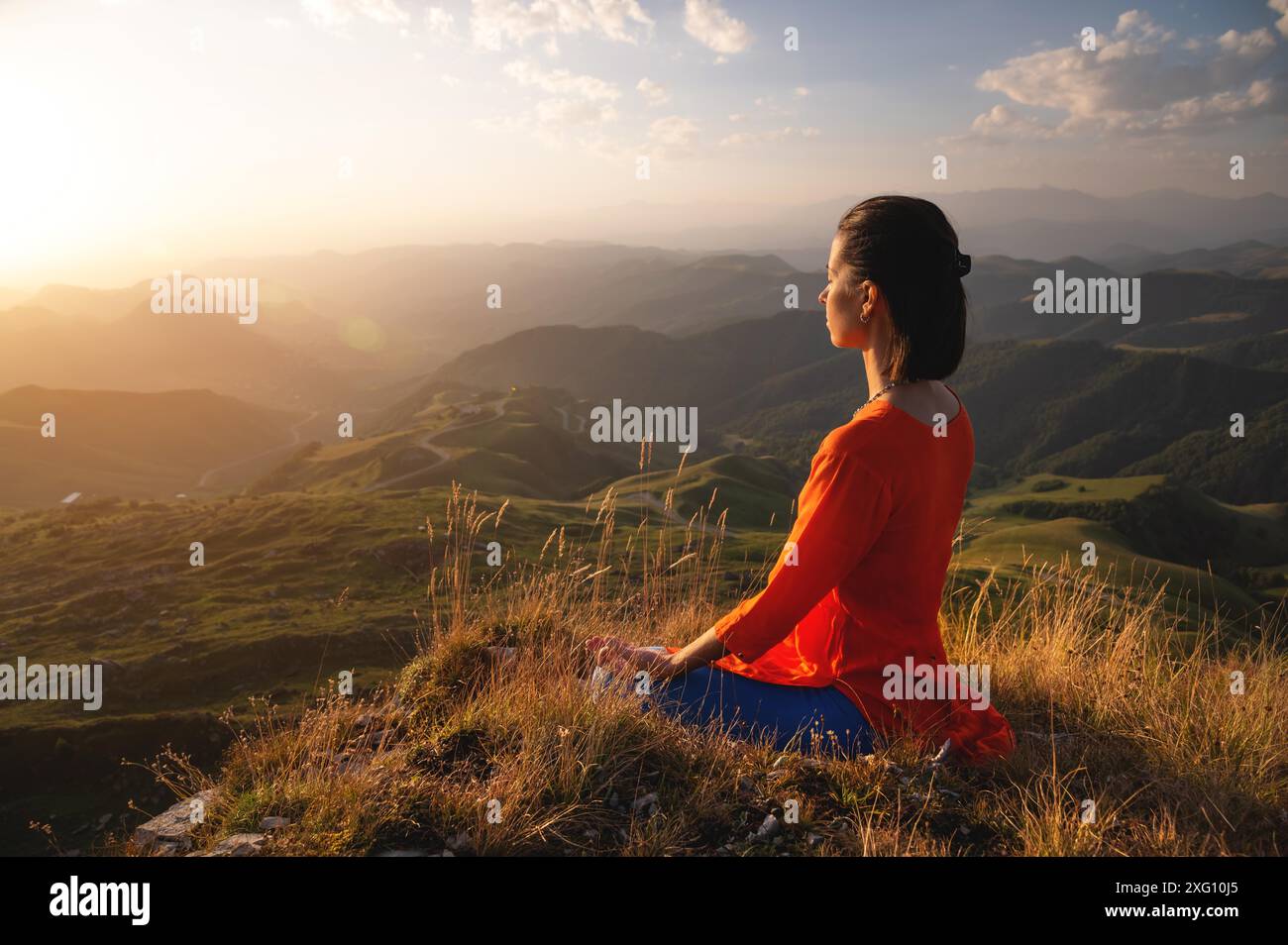 Jeune femme caucasienne médite dans la position du lotus dans l'herbe dans les montagnes près de la falaise dans le soleil couchant Banque D'Images