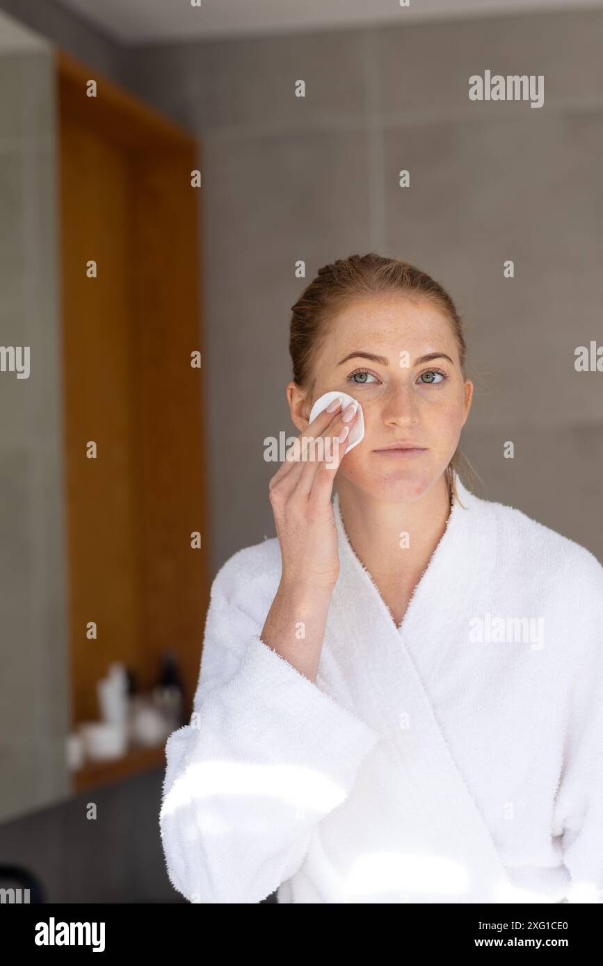 Femme en peignoir nettoyant le visage avec un tampon de coton dans la salle de bain Banque D'Images