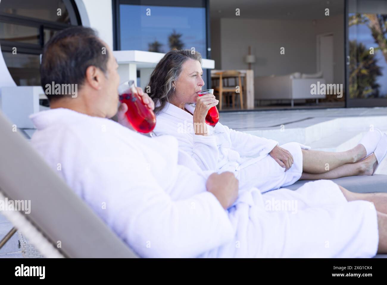 Détente au bord de la piscine, couple senior dans des peignoirs buvant des boissons rouges en vacances Banque D'Images