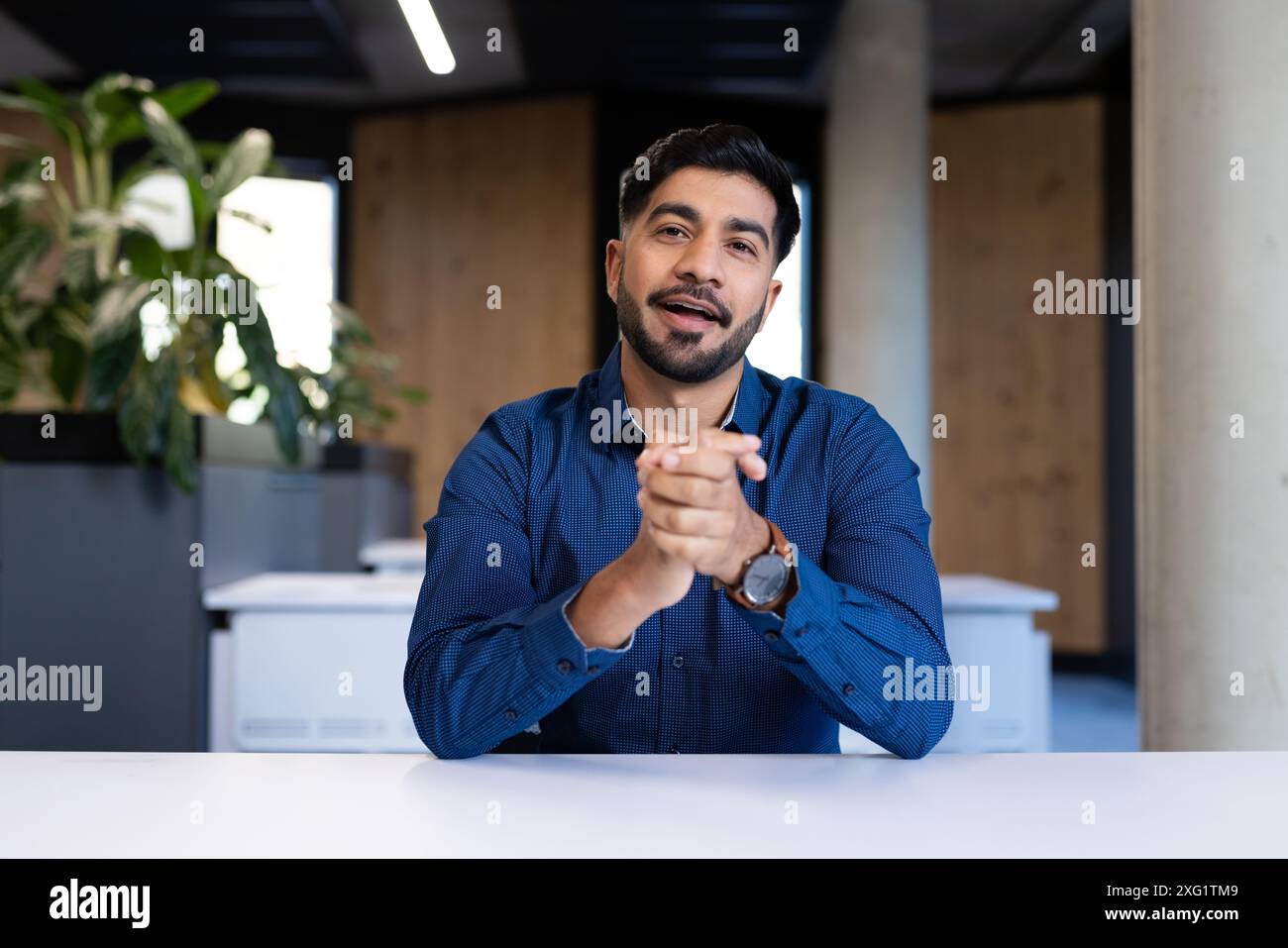 Homme d'affaires en chemise bleue assis au bureau, gestuel et souriant dans le bureau Banque D'Images