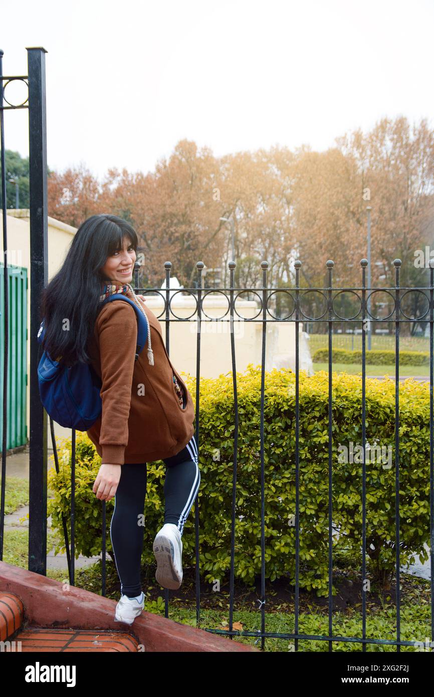 Image verticale de la jeune femme latine drôle dans le parc, elle est debout sur une échelle soutenue par une clôture de sécurité et se tourne pour regarder la caméra souriant, elle est w Banque D'Images