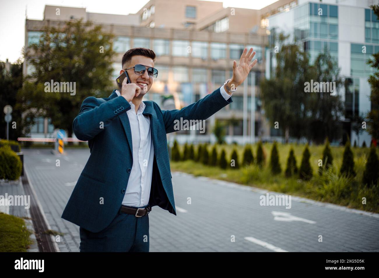 Un jeune homme vêtu d'un costume bleu et de lunettes de soleil se tient dans une rue de la ville parlant au téléphone d'une main tout en faisant signe de l'autre. Banque D'Images