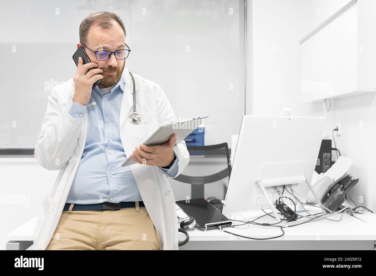 Médecin de sexe masculin avec stéthoscope assis à un bureau, parlant sur un téléphone portable et regardant une tablette numérique, discutant d'un diagnostic de patient. Haute qualité Banque D'Images