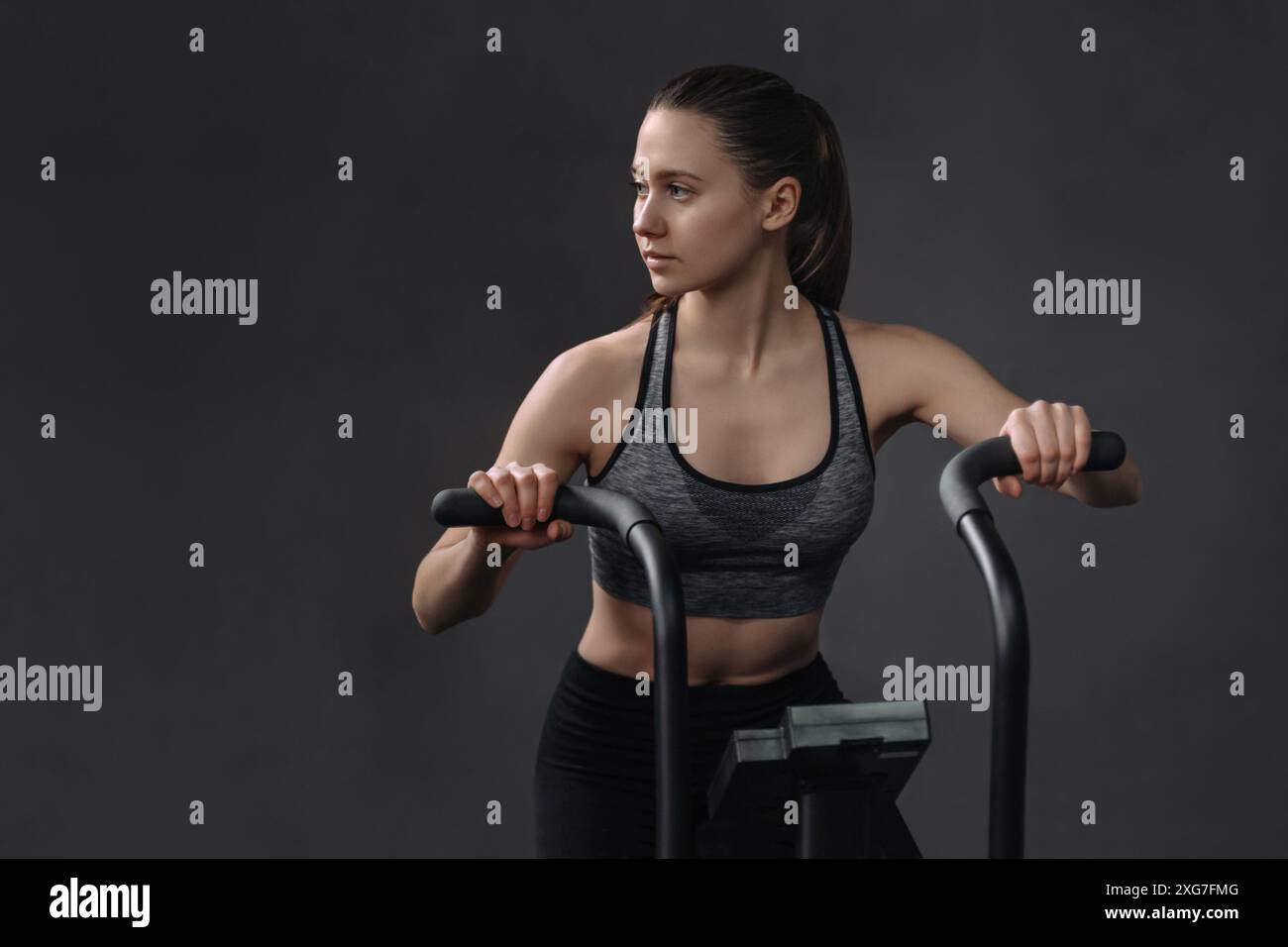 Jeune femme attrayante dans les vêtements de sport en utilisant le vélo d'exercice pour l'entraînement cardio au gymnase de fitness. Fit jolie femelle ayant la classe de vélo d'air. Copier l'espace, isolé sur fond de mur gris Banque D'Images