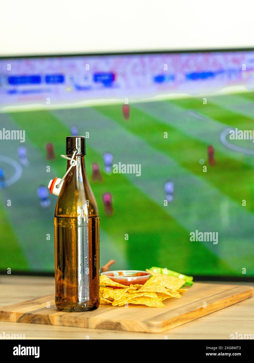 Nachos avec sauce paprika rouge chaude sur un stand en bois, tranches de poivrons ​​green et une boisson contre l'écran de télévision avec un match de football Banque D'Images