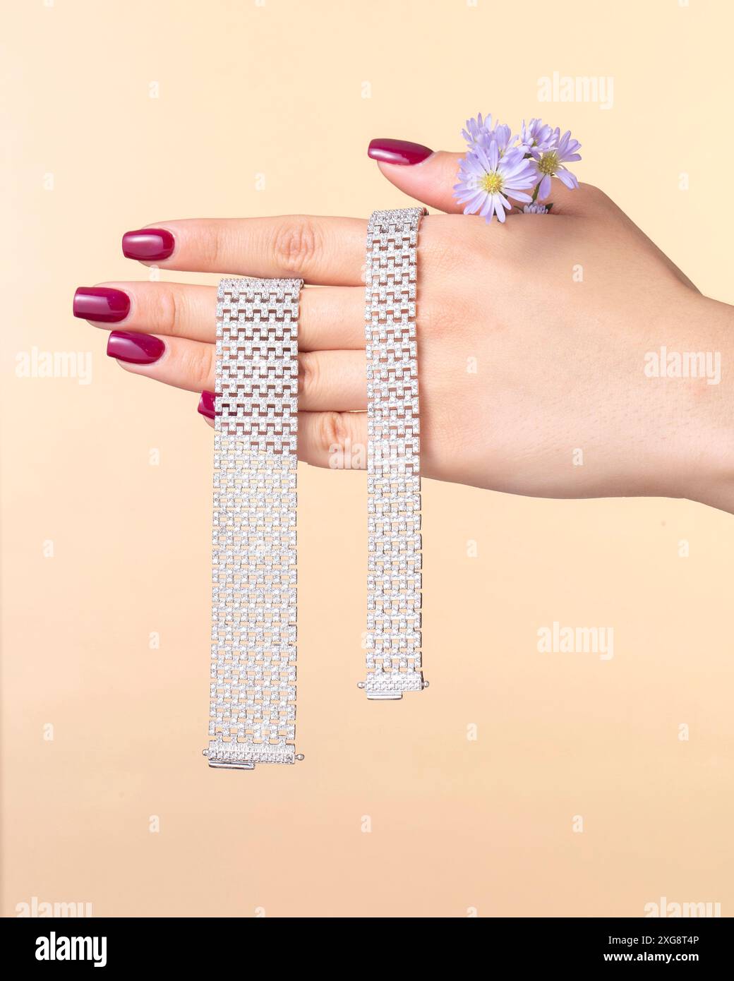 Gros plan d'une main tenant deux bracelets en maille argentée avec une bague florale délicate sur un fond beige doux, soulignant le design élégant. Banque D'Images