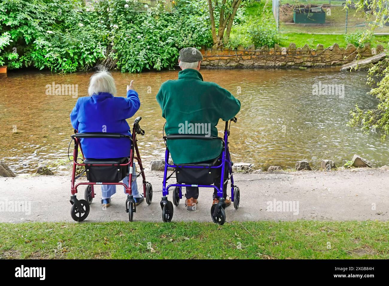 Senior retraité adulte couple d'âge dos vue au repos à côté du ruisseau d'eau Dawlish assis sur des aides à la mobilité le jour froid de juin Devon Angleterre Royaume-Uni Banque D'Images