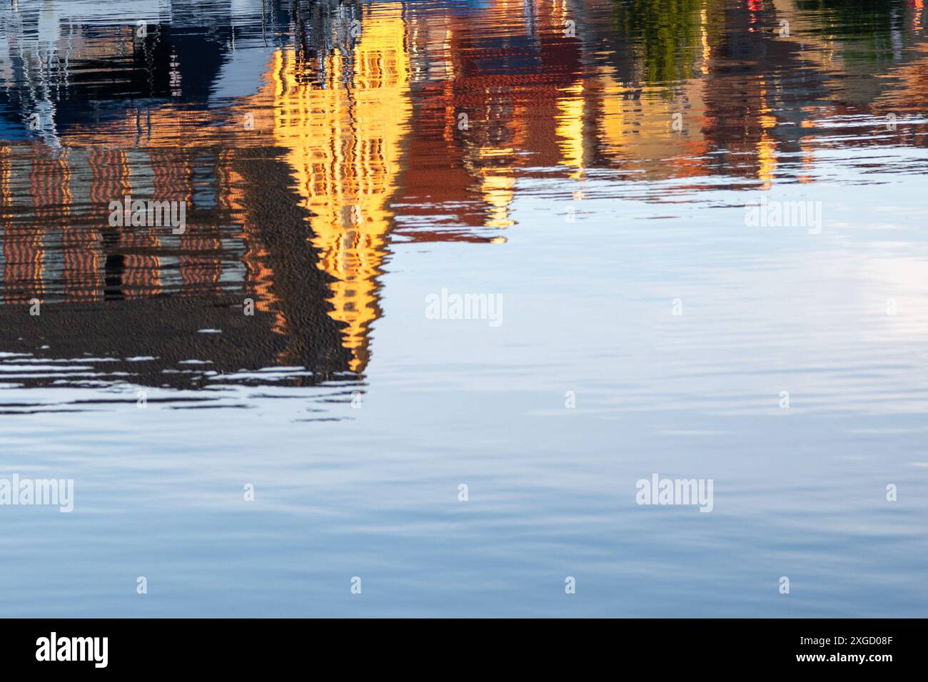 Un plan d'eau calme reflète les teintes jaunes et oranges d'un coucher de soleil, déformant les formes des bâtiments le long du rivage. Banque D'Images