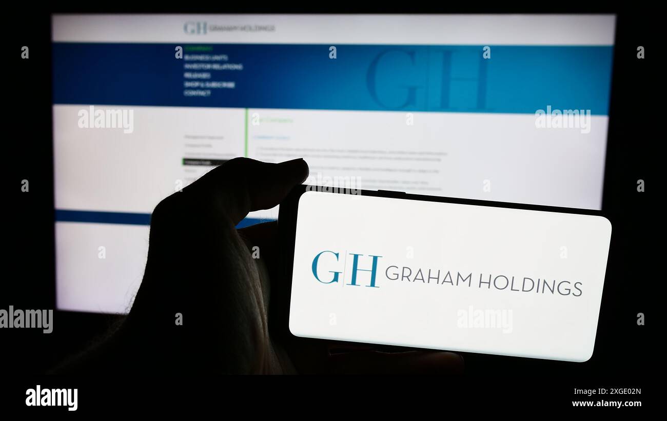 Personne tenant le téléphone portable avec le logo de l'entreprise de médias américaine Graham Holdings Company devant la page Web de l'entreprise. Concentrez-vous sur l'affichage du téléphone. Banque D'Images