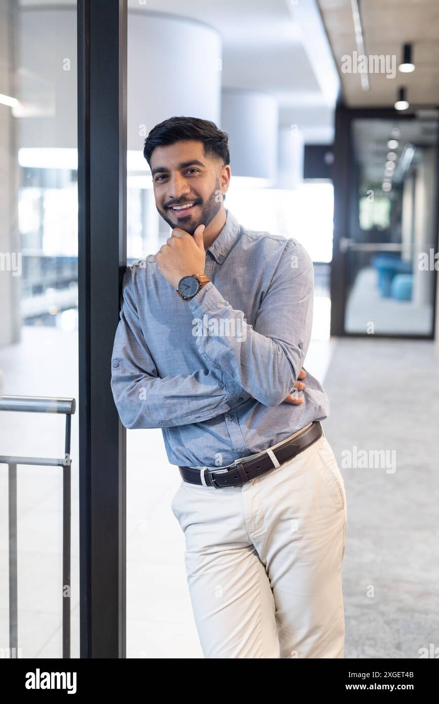 Appuyé contre le mur de verre, souriant homme d'affaires dans le bureau portant une tenue décontractée élégante Banque D'Images
