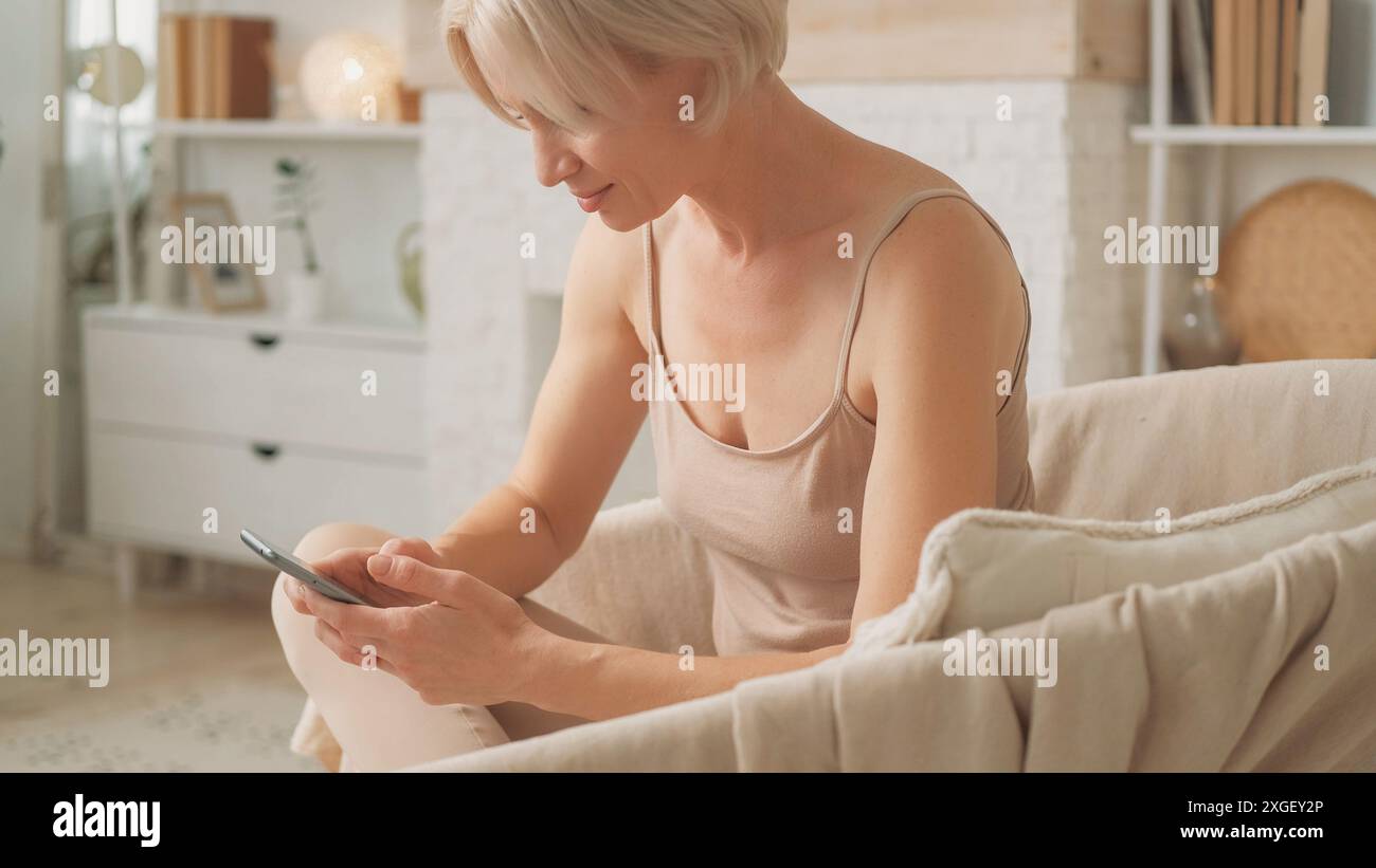 Chat mobile. Communication Internet. Femme heureuse tapant un message sur smartphone gadget profitant de la technologie moderne assise sur le canapé dans l'intérieur du salon lumineux à la maison. Banque D'Images