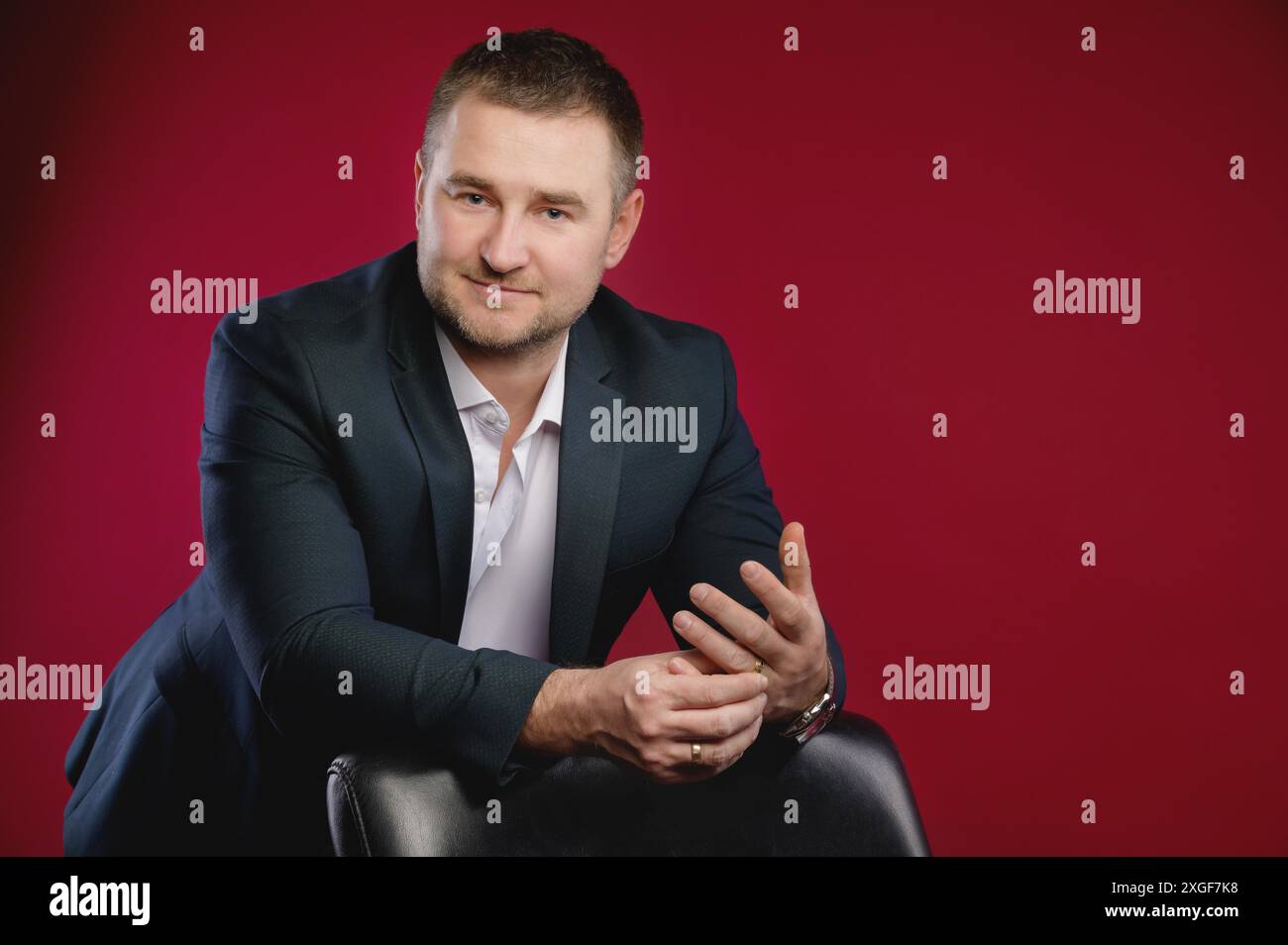 Portrait d'un homme d'affaires caucasien souriant attrayant dans un costume. Se tient dans le studio sur un fond rouge, appuyé sur un fauteuil en cuir et Banque D'Images