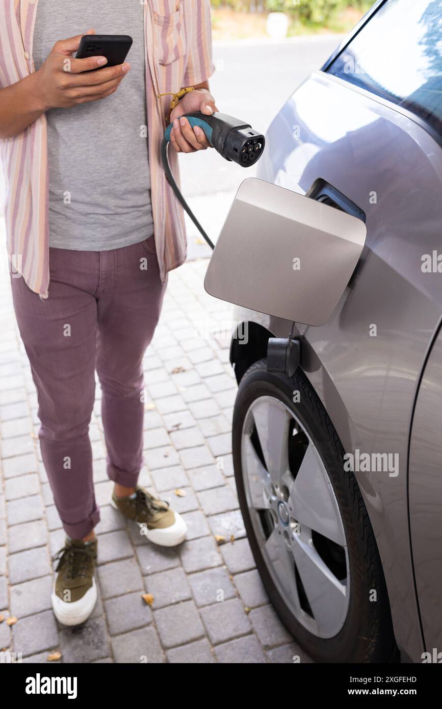 Voiture électrique de charge, homme tenant smartphone et câble de charge dans l'allée Banque D'Images