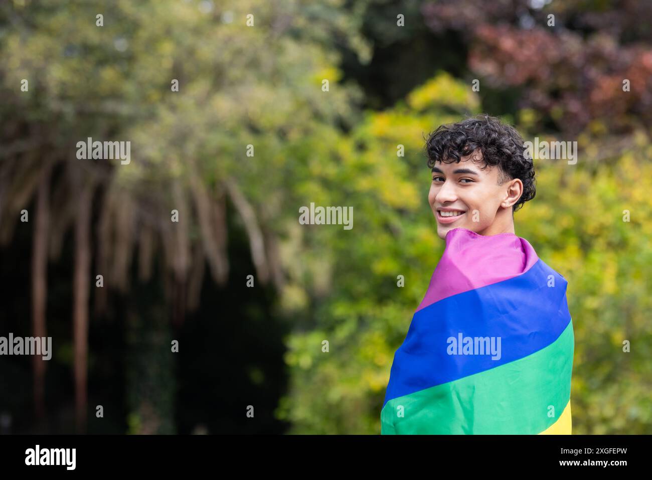 Jeune homme souriant enveloppé dans un drapeau arc-en-ciel, célébrant la fierté LGBTQ+ en plein air, espace de copie Banque D'Images