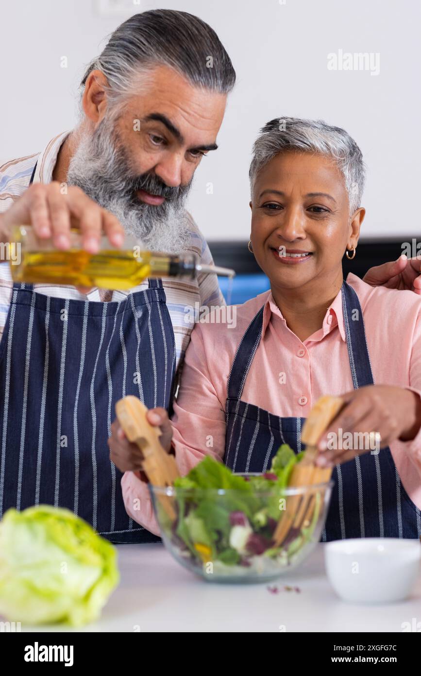 Couple senior préparant la salade ensemble, versant l'huile d'olive et mélangeant les ingrédients Banque D'Images