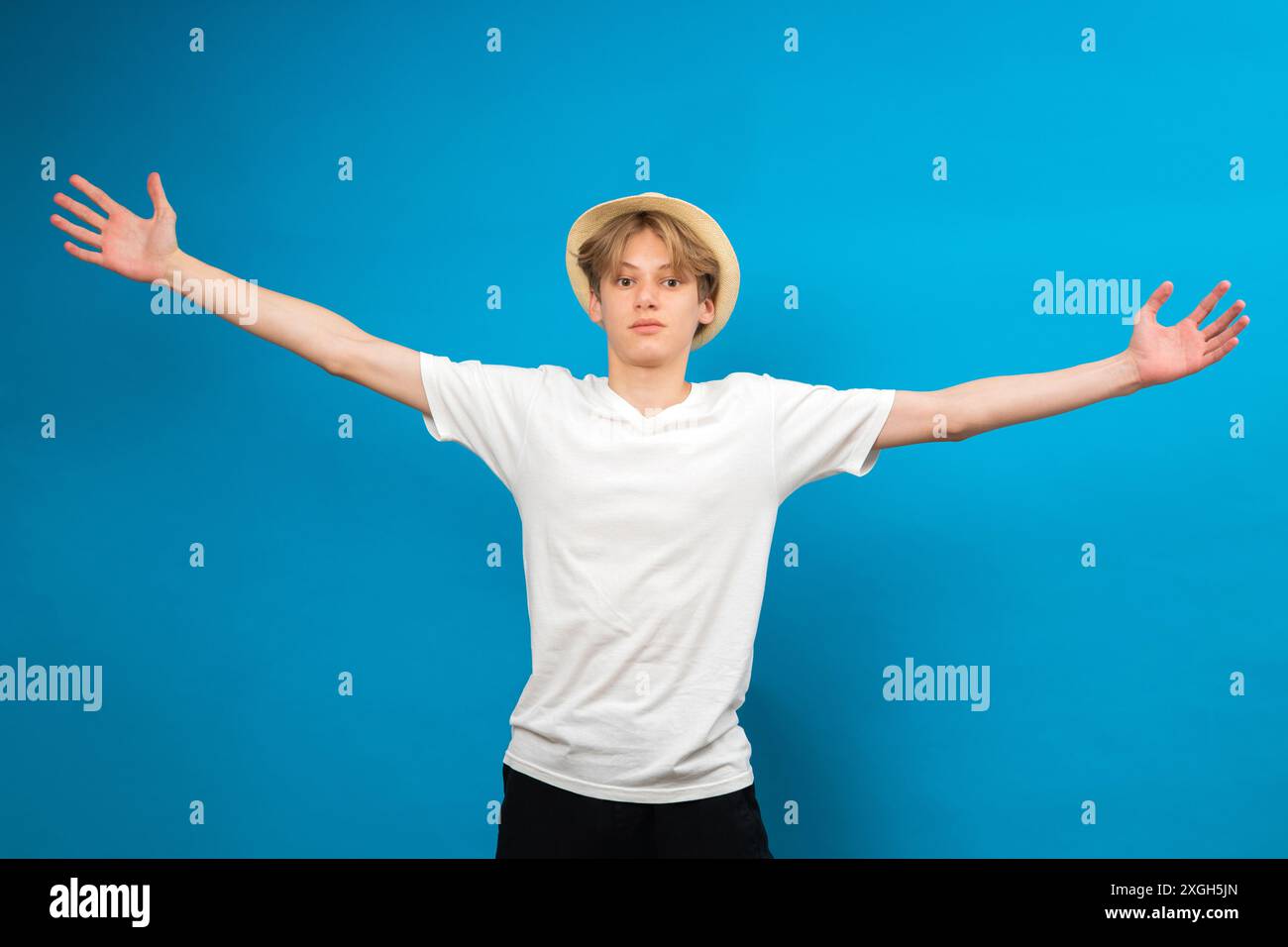 Heureux garçon blond en t-shirt blanc et chapeau sur un fond bleu. Un adolescent dans des vêtements confortables et élégants, a vomi les mains et s'amuse dans le studio Banque D'Images