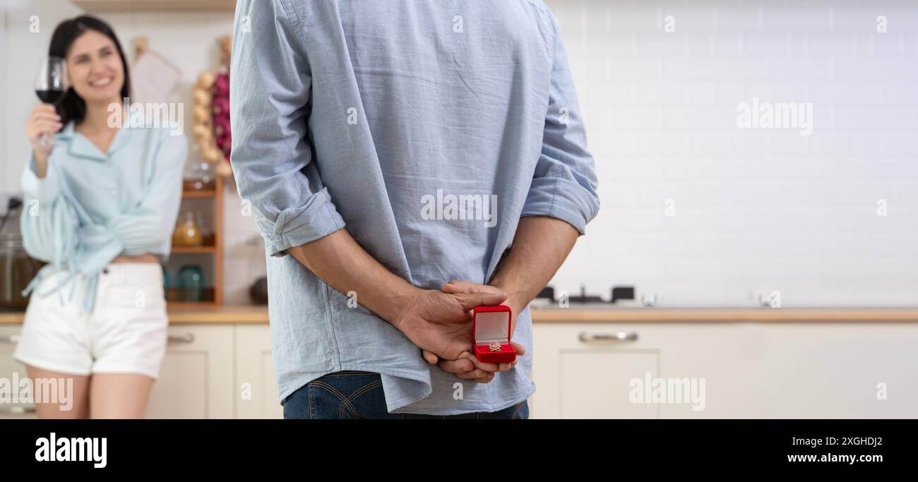 Couple multiethnique ayant un dîner romantique dans la cuisine à la maison. Un homme se tient debout avec une boîte avec une bague de mariage derrière lui, l'offrant à une femme qui me sourit Banque D'Images