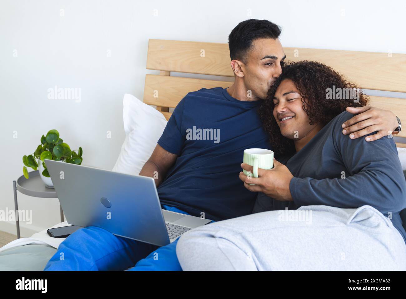 L'homme du moyen-Orient embrasse une jeune femme biraciale dans un cadre de chambre confortable. Ils partagent un moment de détente avec un ordinateur portable et un café, symbolisant le h moderne Banque D'Images