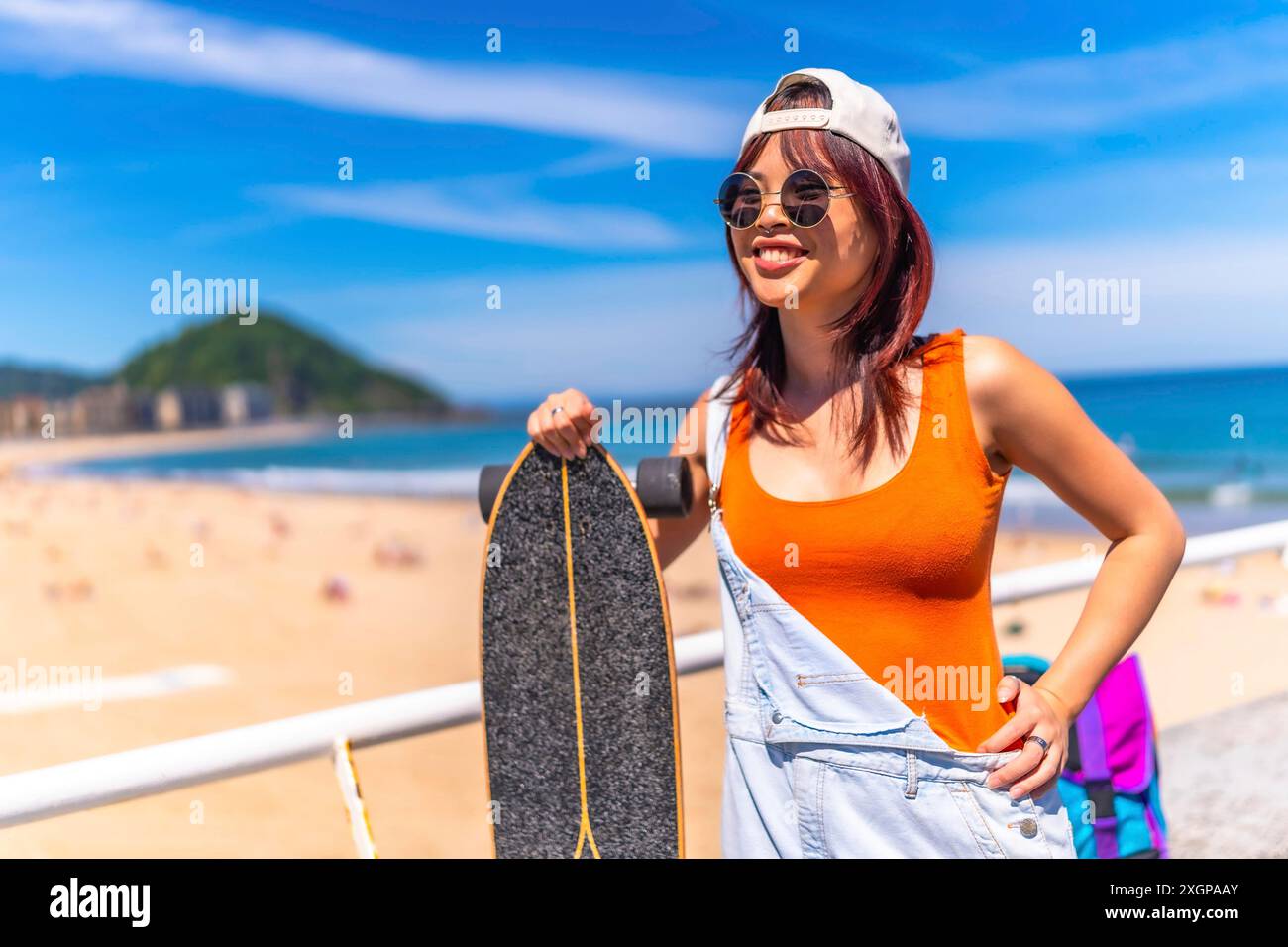 Portrait de trois quarts de longueur d'une jeune femme cool chinoise avec skateboard souriant debout à côté de la plage Banque D'Images