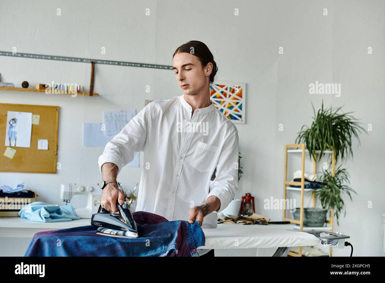 Un jeune homme en chemise blanche repasse soigneusement un morceau de tissu dans son atelier à domicile. Banque D'Images