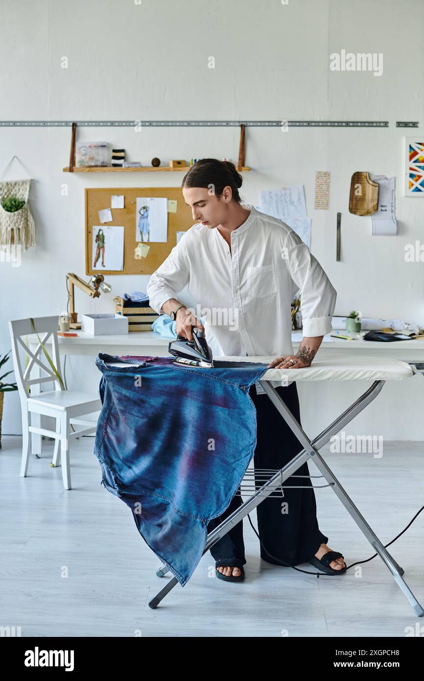 Un jeune homme en chemise blanche repasse un vêtement bleu dans son atelier de restauration de vêtements DIY. Banque D'Images