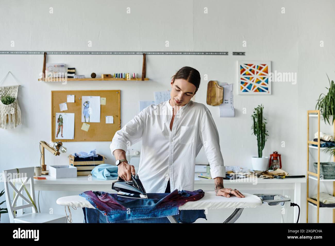 Un jeune homme en chemise blanche travaille sur un projet de restauration de vêtements dans son atelier, en utilisant un fer à repasser pour redonner vie à un vêtement jeté. Banque D'Images
