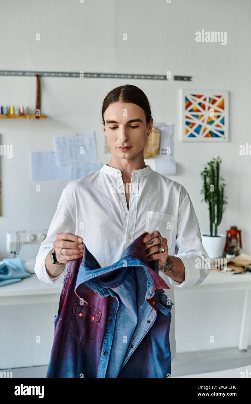 Un jeune homme en chemise blanche examine attentivement un vêtement en denim dans son studio de restauration de vêtements DIY, en se concentrant sur la durabilité en donnant une nouvelle vie Banque D'Images