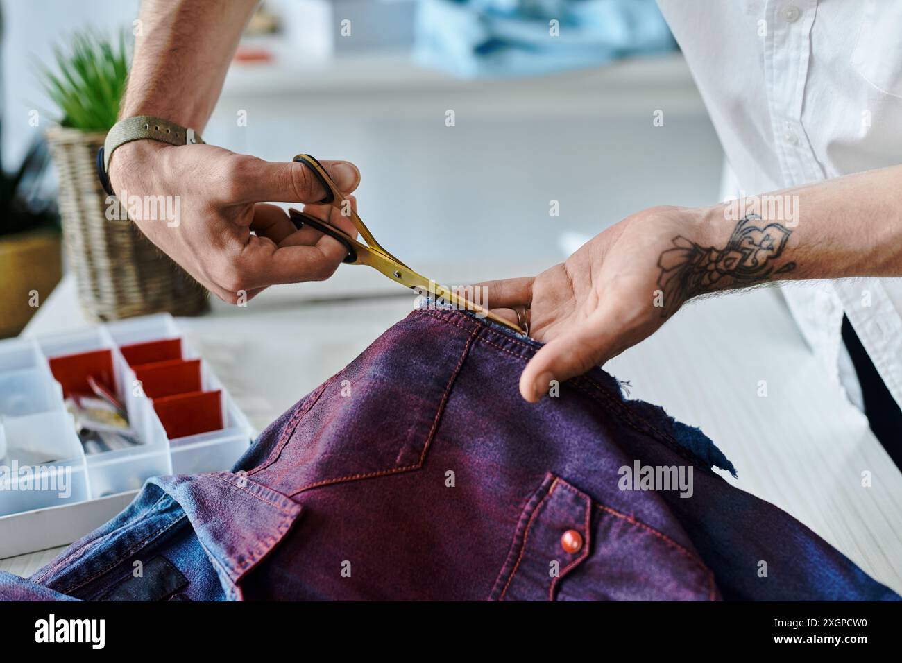 Un jeune homme, coupe soigneusement un morceau de tissu avec des ciseaux dorés dans son atelier, soulignant l'art de l'upcycling et de faire revivre les vieux vêtements. Banque D'Images