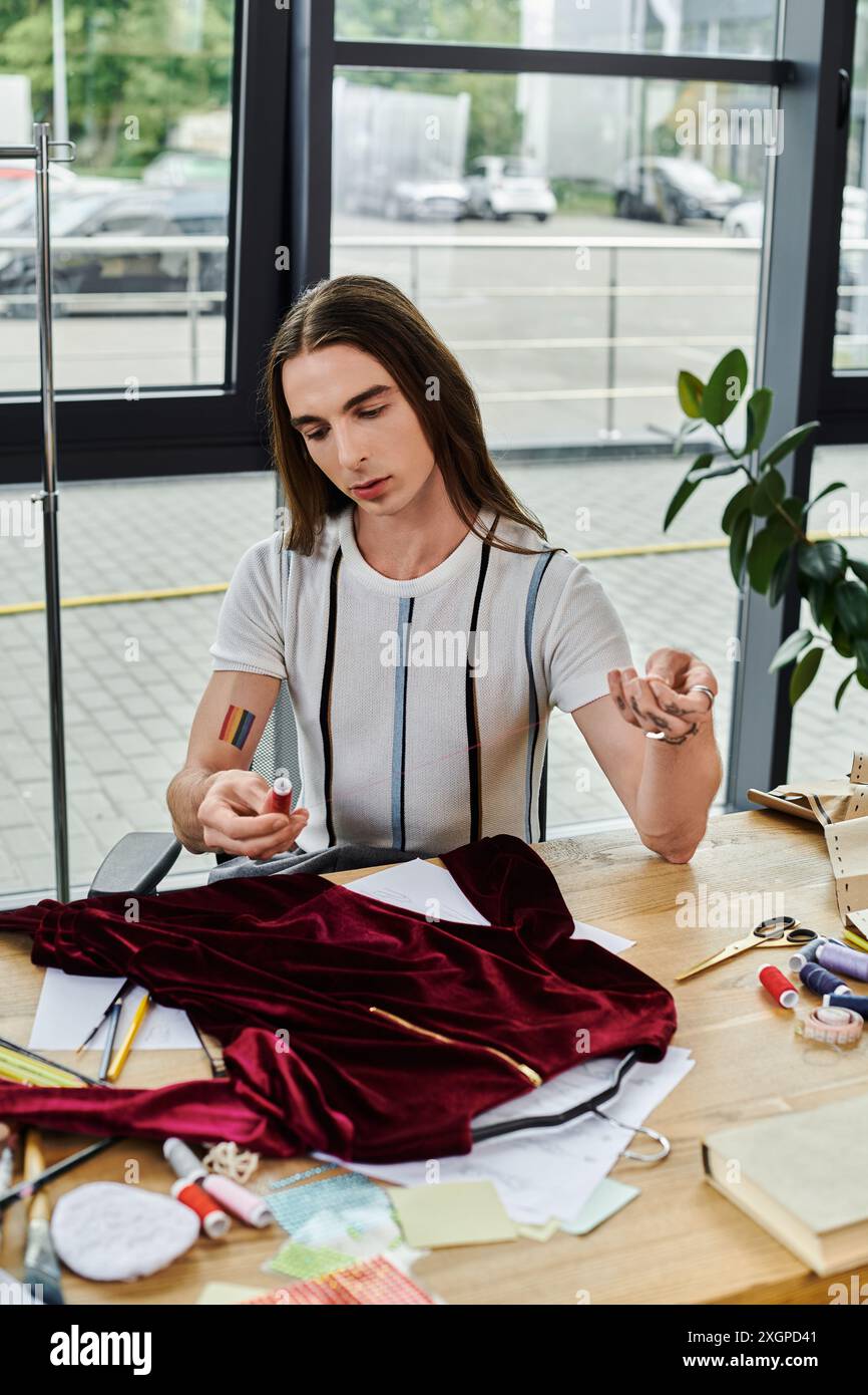 Un jeune homme est assis à son bureau, examinant attentivement une bobine de fil, entouré de débris de tissu et d'outils dans son atelier de restauration de vêtements. Banque D'Images