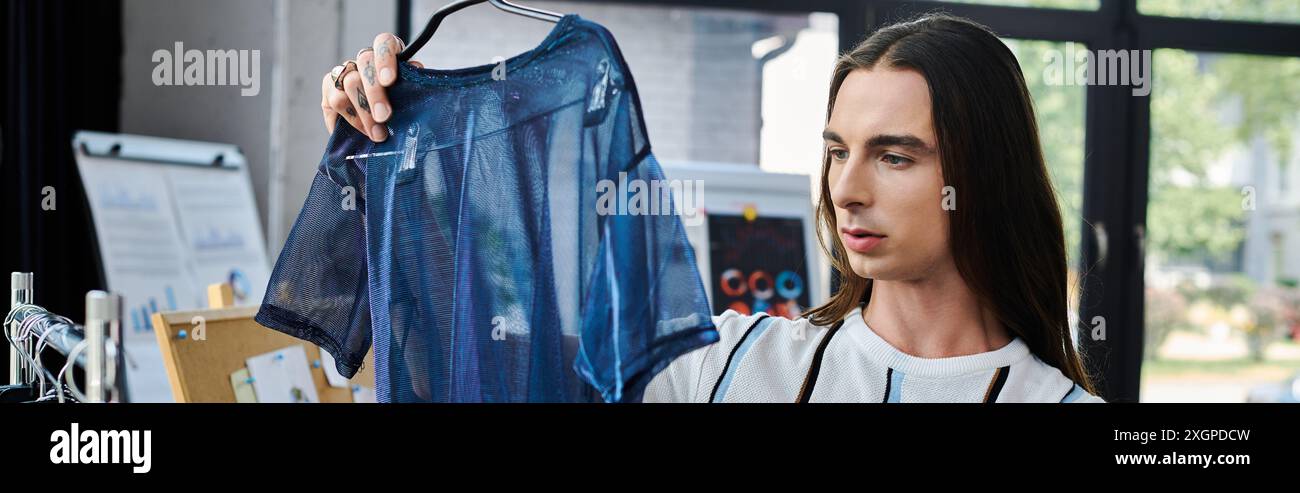 Un jeune homme, concentré intentionnellement, tient un vêtement délicat, bleu, en maille, soulignant sa passion pour la renaissance des vieux vêtements et la promotion durable f Banque D'Images