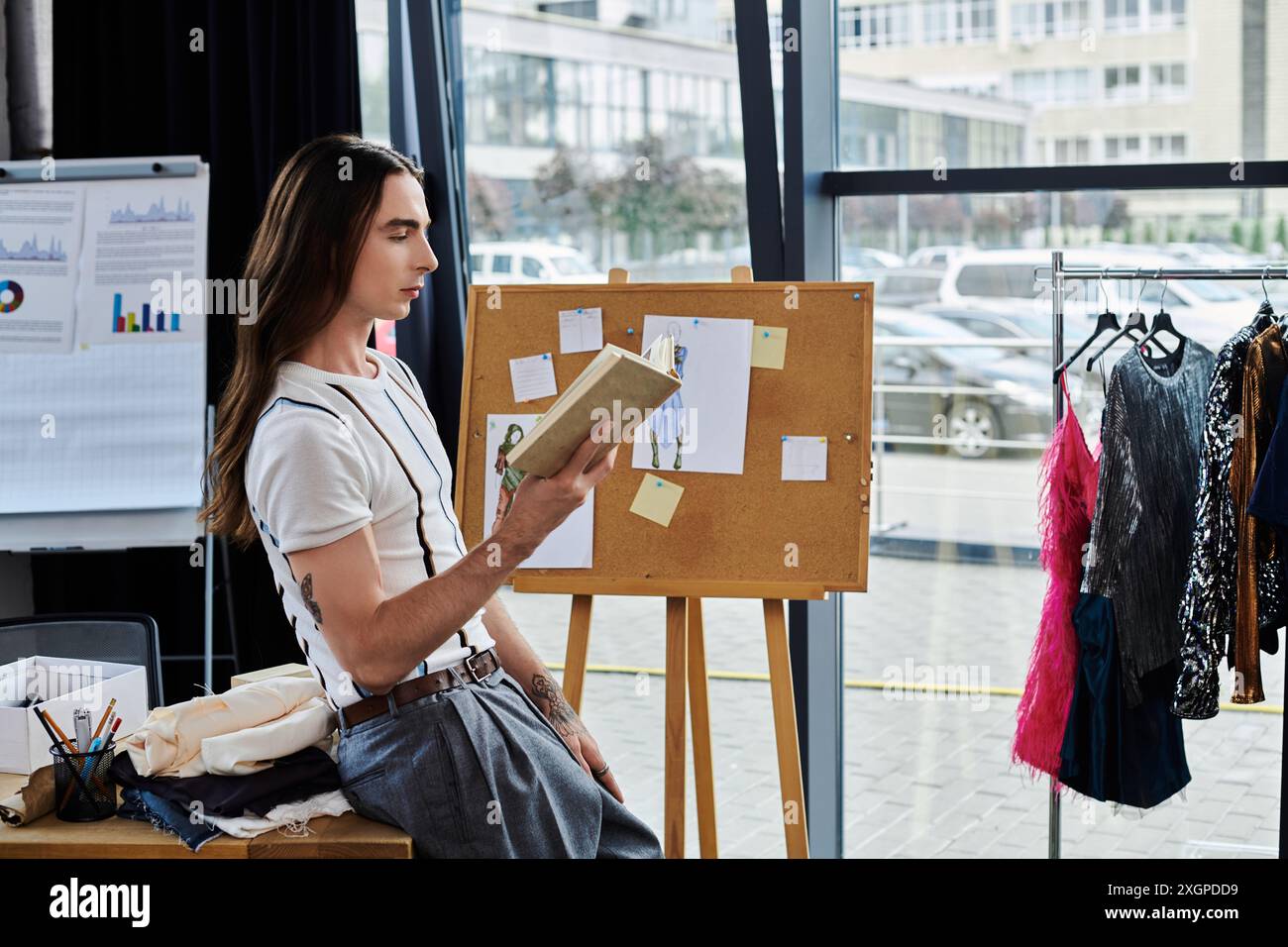Un jeune homme aux cheveux longs est assis dans son atelier de restauration de vêtements, lisant un livre tout en étant entouré par son travail. Banque D'Images