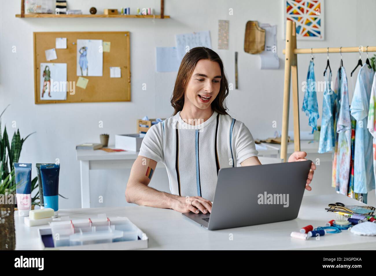 Un jeune homme gay travaille sur son ordinateur portable dans son atelier de restauration de vêtements, entouré de tissus et d’outils. Il se concentre sur donner une nouvelle vie aux vieux vêtements. Banque D'Images