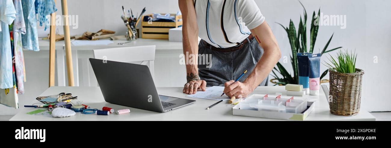 Un jeune homme travaille sur un design dans son atelier de restauration de vêtements DIY, entouré de matériaux et d’outils pour donner une nouvelle vie aux vieux vêtements. Banque D'Images