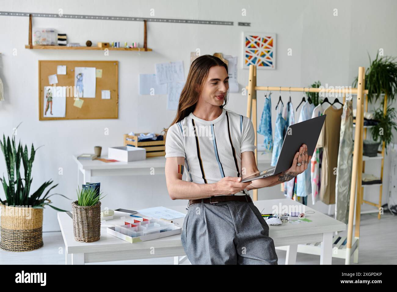 Un jeune homme travaille sur son ordinateur portable dans son atelier de restauration de vêtements, entouré de tissus et de plantes vibrants. Banque D'Images
