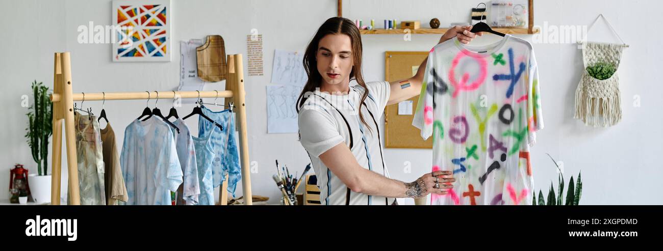 Un jeune homme se tient dans son atelier, affichant fièrement un T-shirt vibrant peint à la main. Il adopte des pratiques durables, insufflant une nouvelle vie à l'ancien Banque D'Images