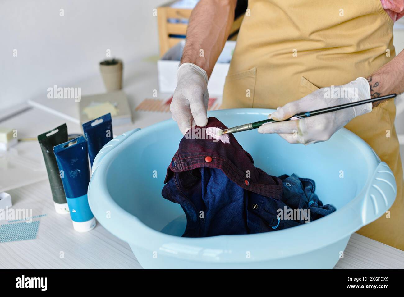 Un jeune homme peint une chemise jetée avec un pinceau dans un bassin d’eau, la revitalisant dans son atelier de restauration de vêtements DIY. Banque D'Images