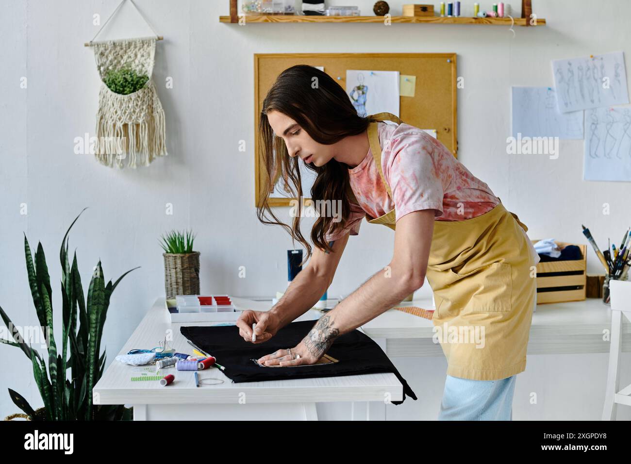 Un jeune homme, aux cheveux longs et à la chemise teintée, travaille sur un vêtement dans son home studio, axé sur la redynamisation des objets jetés. Banque D'Images