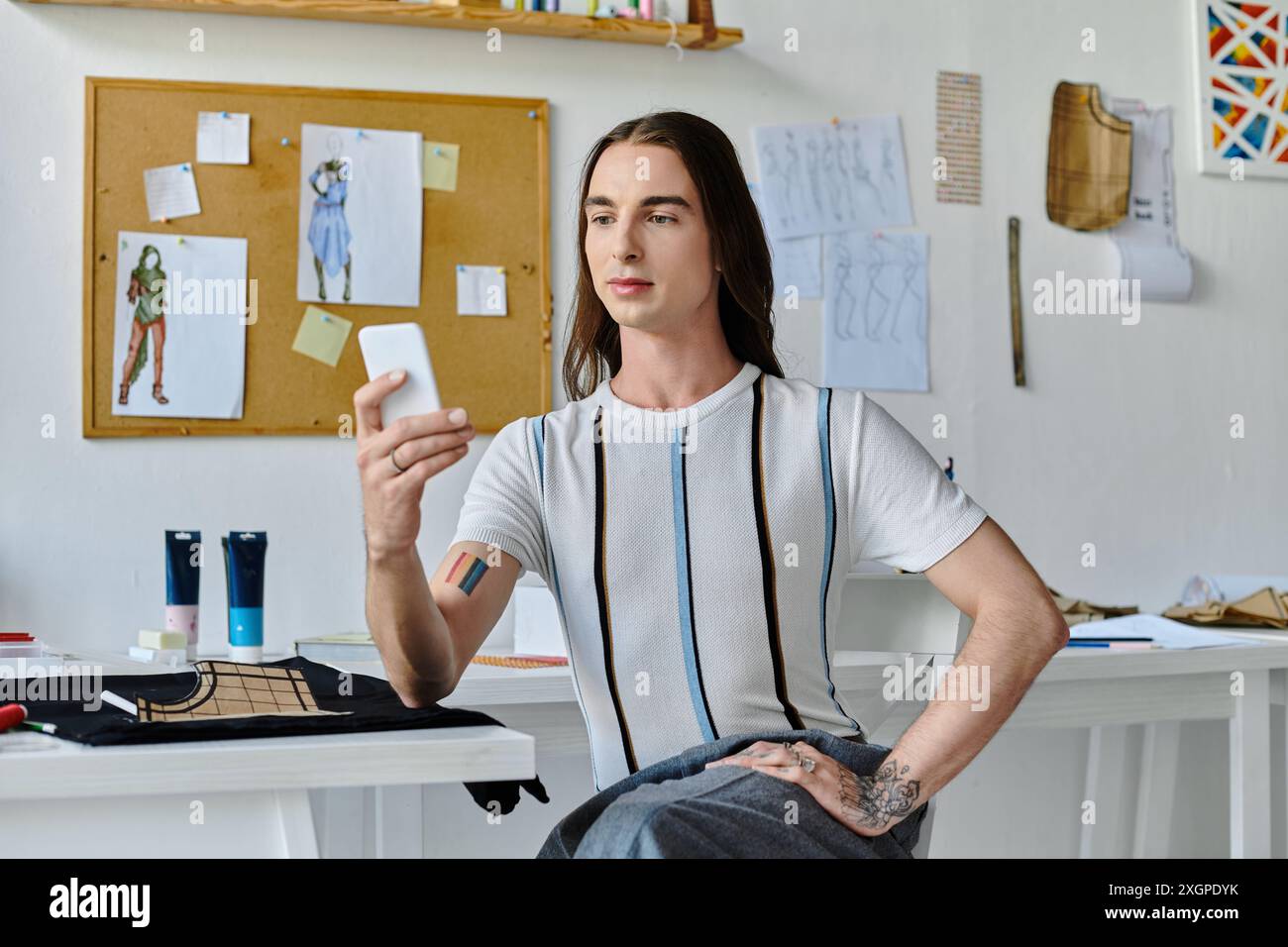 Un jeune homme, assis dans son atelier de restauration de vêtements de bricolage, regarde son téléphone tout en étant entouré de croquis de design et de fournitures. Banque D'Images