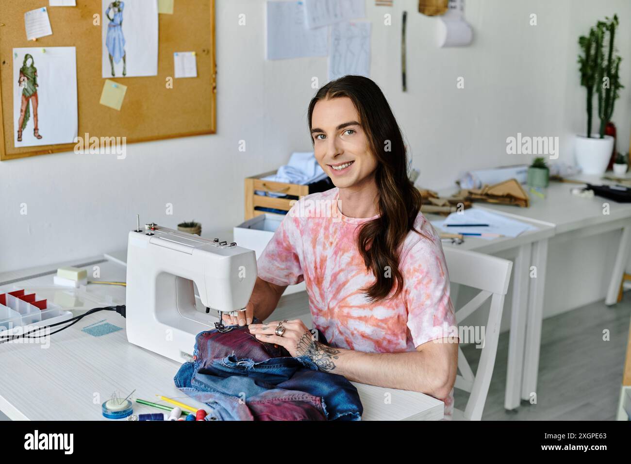 Un jeune homme, vêtu d’une chemise teintée, sourit en travaillant sur une machine à coudre dans son atelier de restauration de vêtements. Banque D'Images