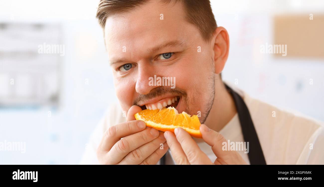 Portrait de Chef professionnel Bite Orange Slice. Homme Vlogger mangeant jus Citrus avec sourire sur le visage. Personne a mis des fruits jaunes à la bouche. Des vies alimentaires saines Banque D'Images