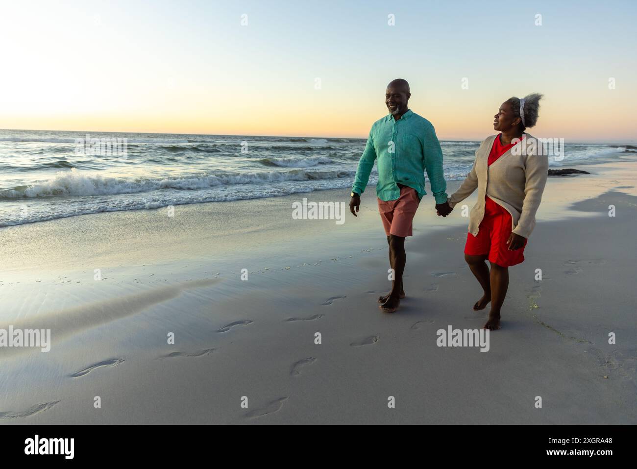 Couple senior afro-américain profite d'une promenade sur la plage au coucher du soleil. Leur promenade détendue le long du rivage capture un moment de retraite sereine. Banque D'Images