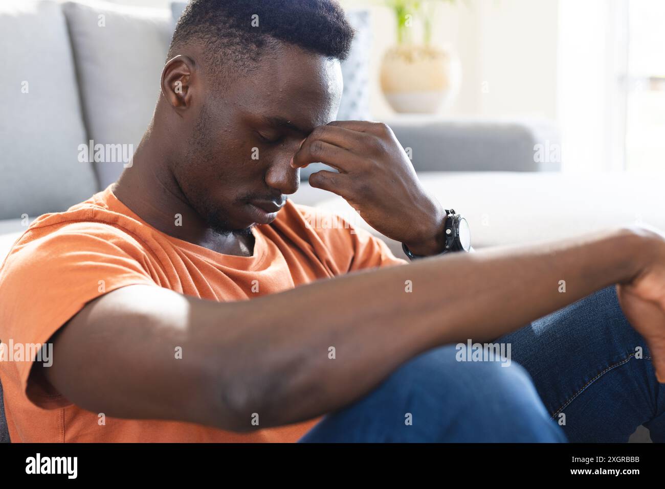 Homme afro-américain réfléchi assis sur le sol dans le salon ensoleillé tenant le nez. Santé mentale, dépression, solitude, émotion, vie domestique et Banque D'Images