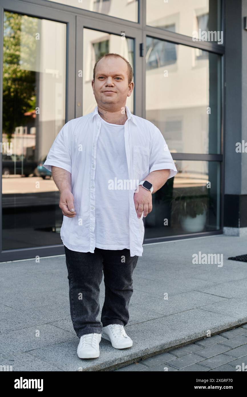 Un homme avec inclusivité dans une chemise blanche marche sur un trottoir à l'extérieur d'un bâtiment avec de grandes portes vitrées. Banque D'Images