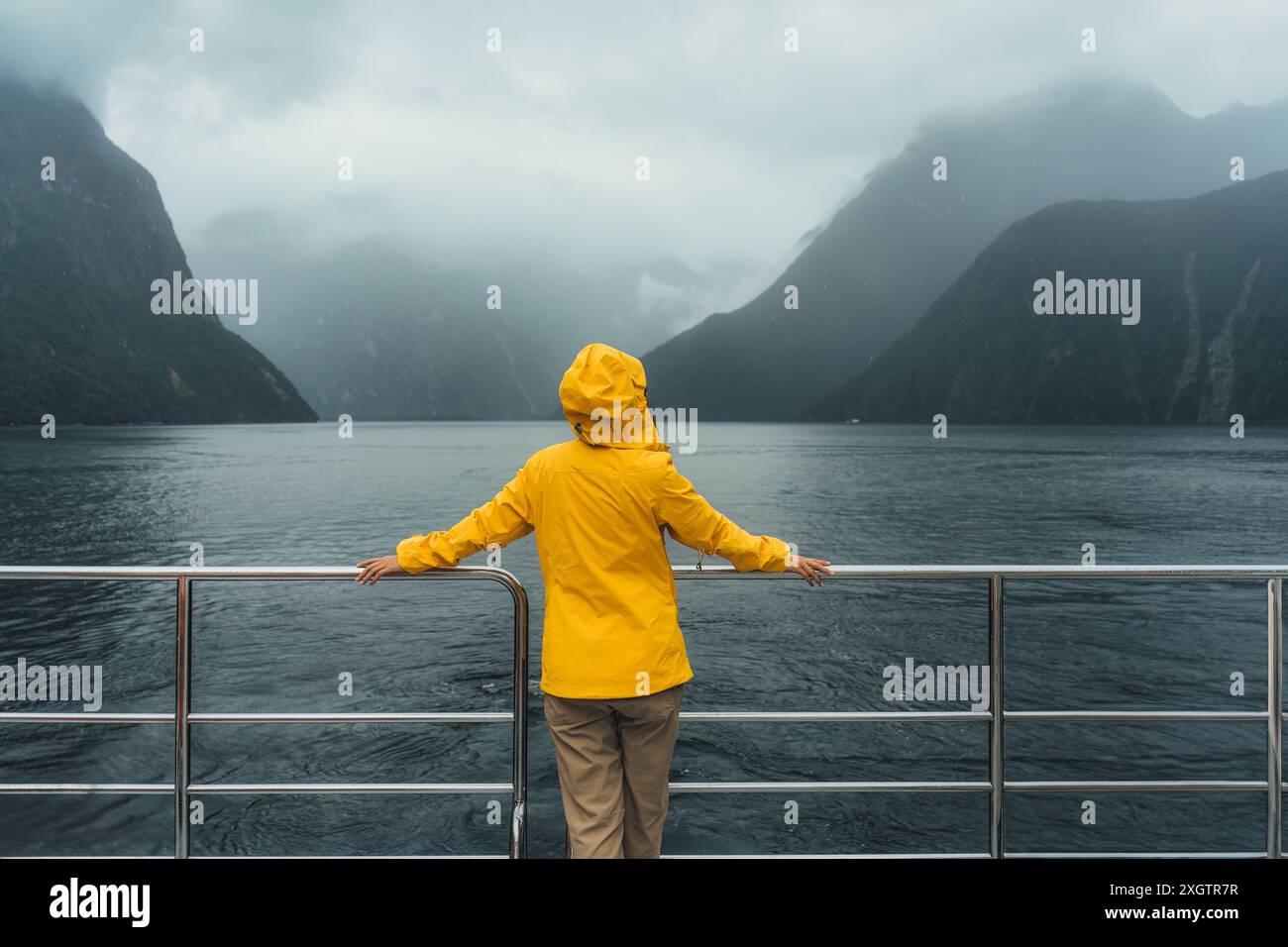 Vue arrière d'une jeune femme en veste jaune profitant de la vue de Milford Sound pendant la croisière en ferry en vacances au parc national de Fjordland, Nouvelle-Zélande Banque D'Images
