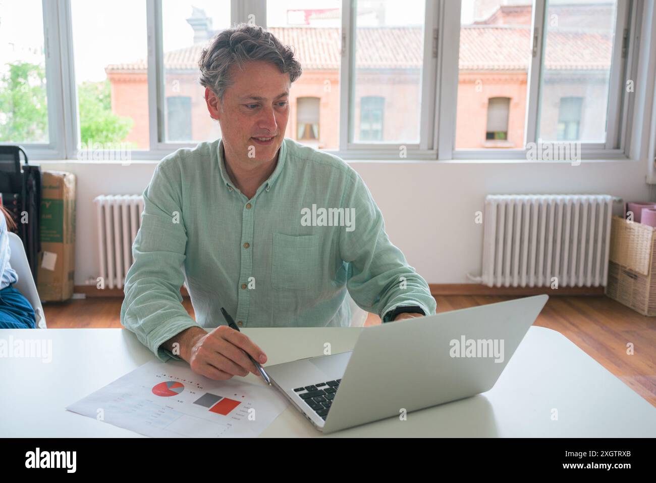 Un homme professionnel mature évalue les données sur un ordinateur portable, note des notes dans un bureau ensoleillé, mettant en valeur une scène commerciale typique Banque D'Images