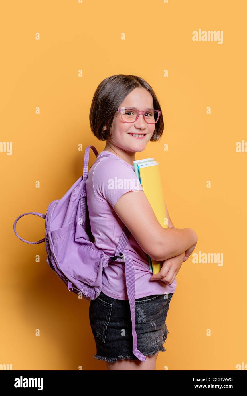 Une jeune fille joyeuse avec des lunettes, tenant des livres et portant un sac à dos violet, sourit en regardant l'appareil photo sur un fond jaune vif Banque D'Images