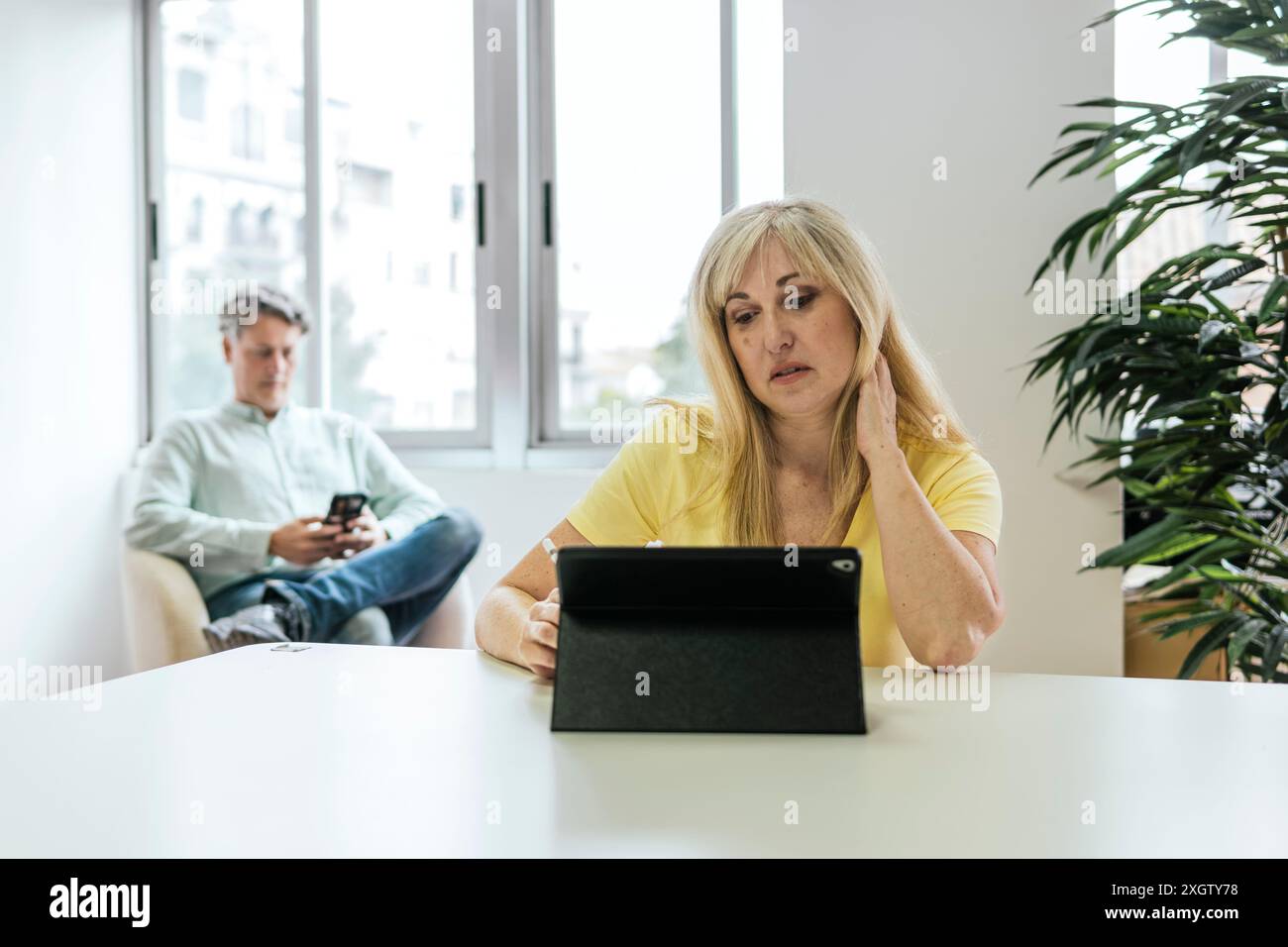 Une femme caucasienne regarde sa tablette dans un espace de coworking tandis qu'un homme caucasien utilise son smartphone en arrière-plan, illustrant ainsi le travail moderne Banque D'Images